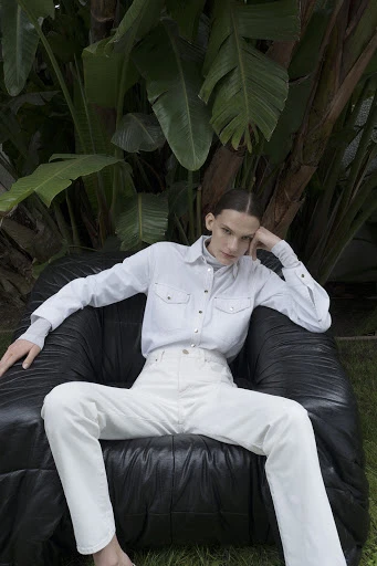 A fair-skinned adult in white pants and shirt, posing in a black leather chair surrounded by tropical plants in an outdoor environment.