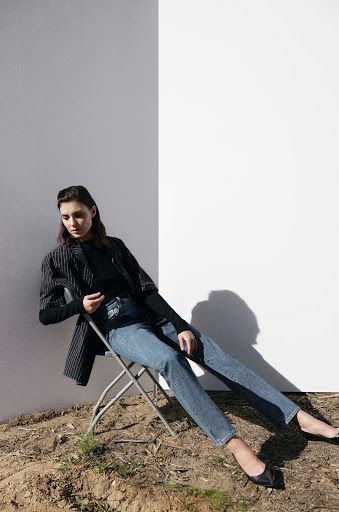 A young adult woman sits on a folding chair outdoors wearing blue jeans, black top and blazer against a two-tone white and grey wall.