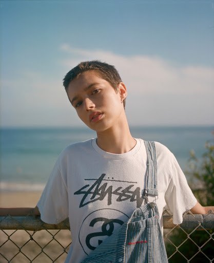 Young woman in Stussy tee shirt and overalls posing outdoors against a beach and blue sky backdrop. Fashion editorial style.