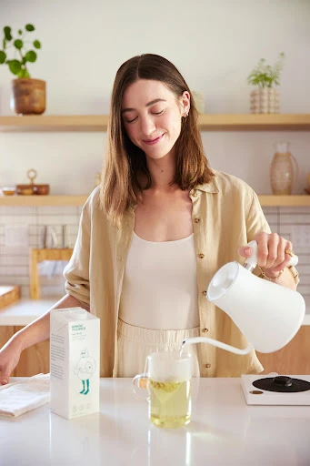 Woman pouring liquid from white pitcher into glass mug with chamomile tea on a bright kitchen counter, next to plant-based milk.