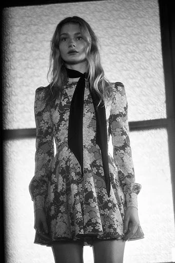 Monochrome portrait of a fair-skinned adult woman with long blonde hair, wearing a floral dress and black neck scarf, looking at camera.