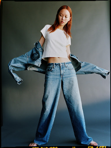 Model with red hair wearing a white t-shirt and loose denim jeans holding out a denim jacket in a studio.