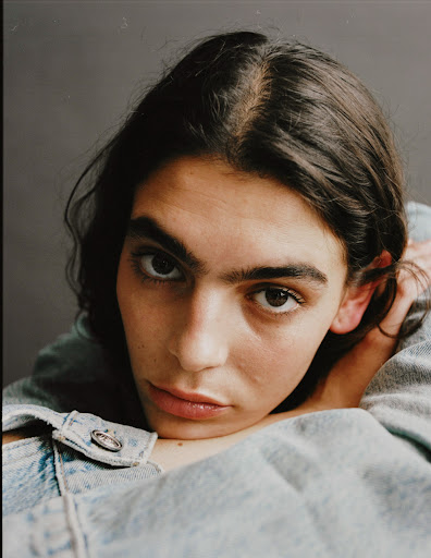Close-up portrait of a young adult woman with dark hair, wearing a light-wash denim jacket, against a solid gray background in a studio setting.
