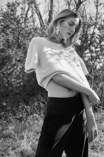 A woman in a cream-colored sweater and dark pants poses outdoors in a black and white medium shot with natural lighting.
