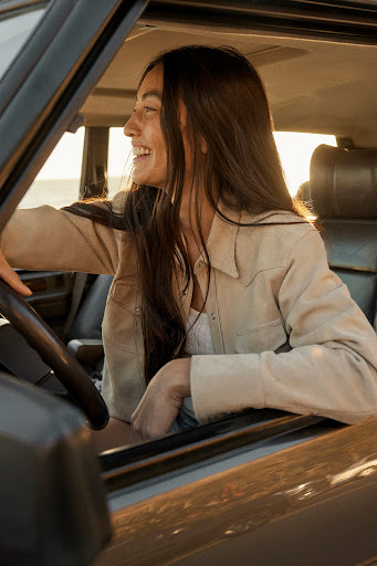 A woman smiles in the driver's seat of a classic car on a sunny day. The scene captures a casual, candid moment with a warm, golden light.