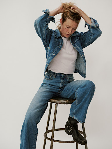 A woman with blonde hair sits on a stool, wearing a denim jacket, jeans, a t-shirt and black boots, against a neutral background.