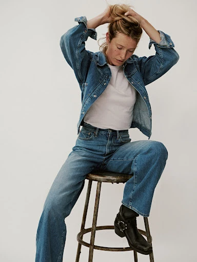 A woman with blonde hair sits on a stool, wearing a denim jacket, jeans, a t-shirt and black boots, against a neutral background.