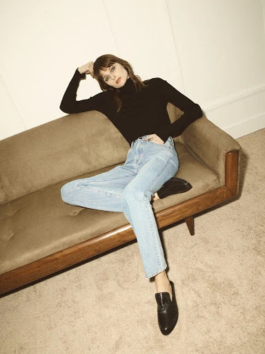 A stylish woman poses on a brown sofa in a fashion shot, wearing a black turtleneck, light blue jeans, and black loafers indoors.