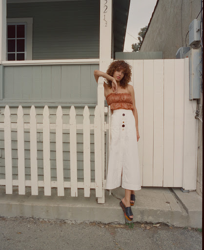 A woman poses in front of a white picket fence wearing a strapless top and white skirt, standing on a residential street with a house.