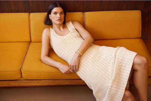 A woman reclines on a mid-century modern style couch in a cream colored dress and gold jewelry indoors in a bright, soft light.