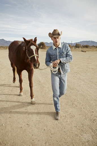 A young adult male wearing a cowboy hat and denim jacket leads a brown horse in a dry, open landscape under a clear sky.