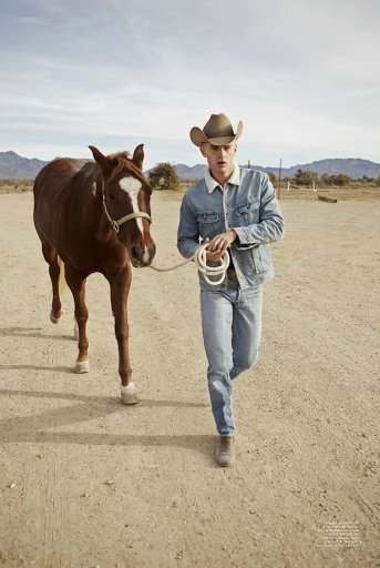 A young adult male wearing a cowboy hat and denim jacket leads a brown horse in a dry, open landscape under a clear sky.