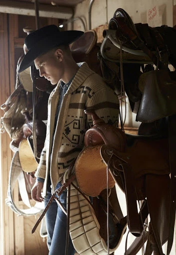 Young man wearing a cowboy hat and patterned cardigan stands in a barn-like space surrounded by several brown leather horse saddles.