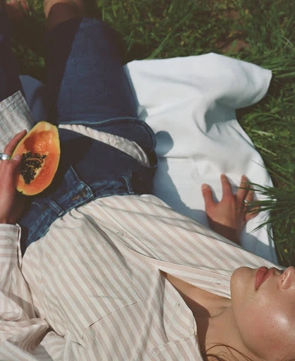 Young woman lounges in a field with a papaya, wearing a striped shirt and jeans, enjoying a peaceful moment in a natural setting.