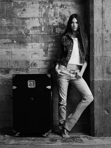 Black and white portrait of a woman in a denim jacket and jeans leaning against a textured wall with a vintage case.