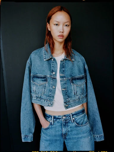 Portrait of a woman in a denim jacket and jeans against a dark background, showcasing a fashion-forward and casual style.
