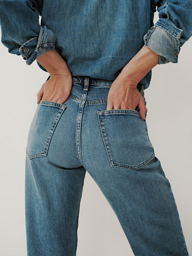 Woman in a denim shirt and jeans, with hands in pockets, posing against a white background in a fashion photography shot.