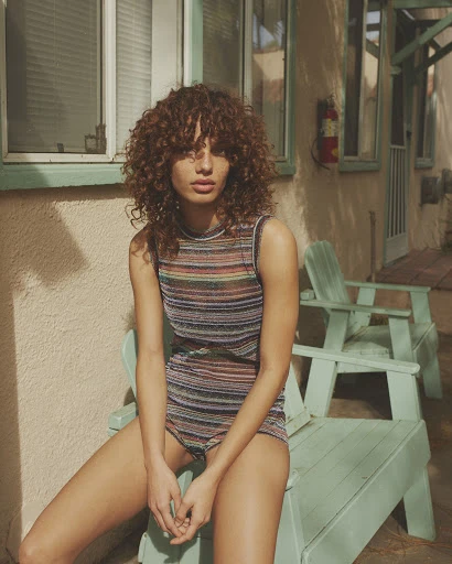 A woman with curly hair is sitting on a blue chair, wearing a striped top and shorts in a casual outdoor setting with natural lighting.
