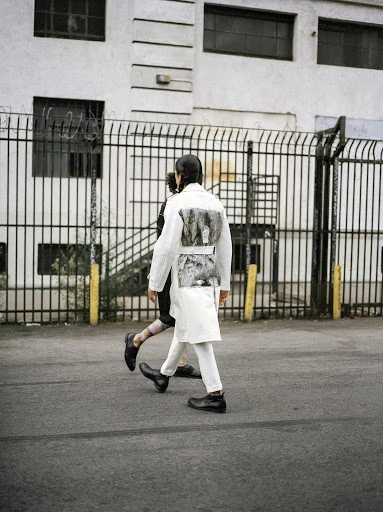 Two people are walking on an asphalt street, one wearing a long white jacket and pants, with a building and fence in the background.