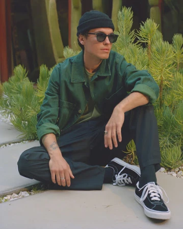 A young man wearing a green jacket, black beanie, and sunglasses sitting outdoors near cacti.