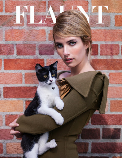 Emma Roberts poses with a black-and-white cat against a brick wall in a studio shot, wearing a tan coat and smokey eye makeup.