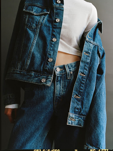 Denim jacket and jeans outfit on a human in a studio. The jacket is casually draped over the shoulder. The background is a plain, grey color.