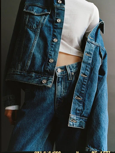 Denim jacket and jeans outfit on a human in a studio. The jacket is casually draped over the shoulder. The background is a plain, grey color.