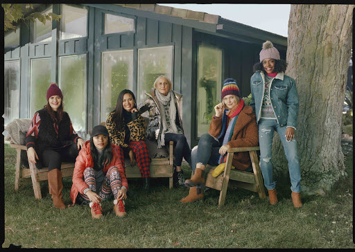Group of six women in winter clothes pose outdoors in front of a green building and trees, smiling in a medium shot.