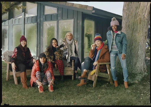 Group of six women in winter clothes pose outdoors in front of a green building and trees, smiling in a medium shot.