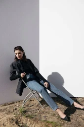 Woman in dark pinstripe blazer and blue jeans seated backward on metal folding chair outdoors against white wall.