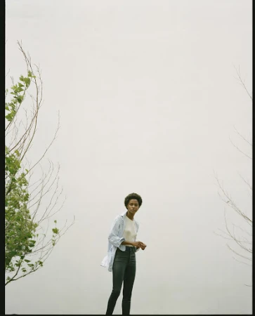 Model standing outdoors wearing a light blue unbuttoned shirt over a white top and dark trousers against a pale overcast sky