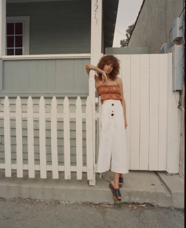Woman with curly brown hair wearing a pleated orange top and long white skirt leaning against a white picket fence.