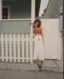 Woman with curly brown hair wearing a pleated orange top and long white skirt leaning against a white picket fence.
