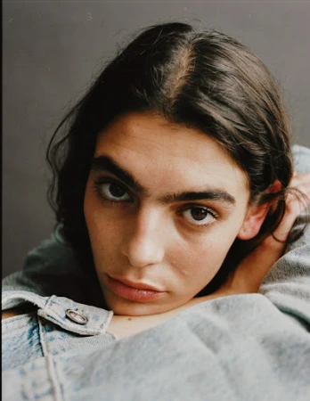 Close-up portrait of a person with long dark hair wearing a light denim jacket against a neutral grey background.