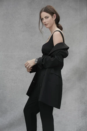 Woman wearing a black blazer draped over her shoulders and matching trousers against a grey textured studio backdrop