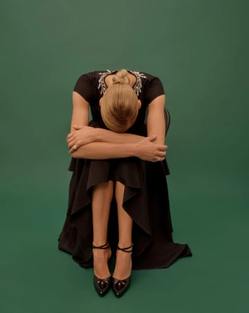 Model seated facing away wearing a black dress and heels against a dark green backdrop styled by Annina Mislin