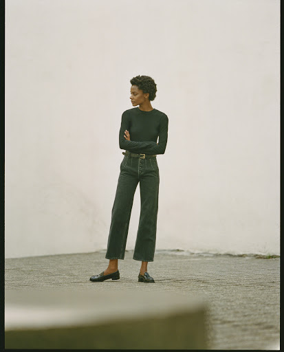 Black woman with short curly hair wearing a dark long-sleeve top and high-waisted dark jeans standing outdoors