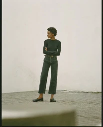 Black woman with short curly hair wearing a dark long-sleeve top and high-waisted dark jeans standing outdoors