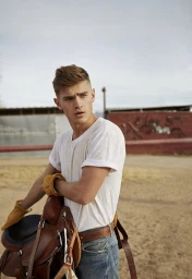 Young man in a white t-shirt and jeans holding a leather saddle in an outdoor sandy setting styled by Annina Mislin.