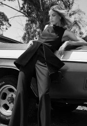 Monochrome fashion photograph of a woman in dark attire leaning against a classic car outdoors.