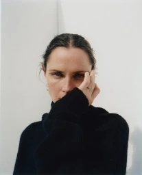 Close-up portrait of a woman in a black turtleneck sweater holding her hand to her face against a white wall.