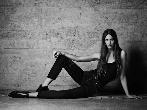 Monochrome fashion portrait of a long-haired woman seated on a concrete floor leaning against a textured brick wall.