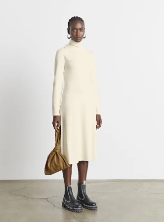 Model wearing cream turtleneck dress and black boots holding a tan handbag against a plain background