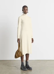 Model wearing cream turtleneck dress and black boots holding a tan handbag against a plain background