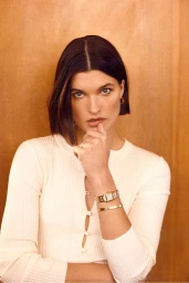 Close-up portrait of a woman with dark bobbed hair wearing a white cardigan and gold jewelry against a warm wood background.