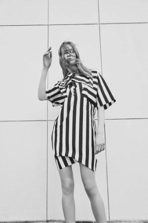Model in a black and white striped dress posing against a gridded wall with hair blowing across her face.