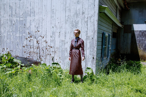 Adult female posing in a printed dress in a verdant, overgrown field beside a weathered building with peeling white paint and blue shutters.