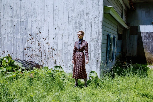 Adult female posing in a printed dress in a verdant, overgrown field beside a weathered building with peeling white paint and blue shutters.