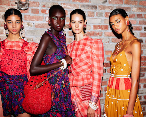 Four fashion models pose in a photo studio wearing colorful dresses. Brick wall background adds texture and depth to the composition.
