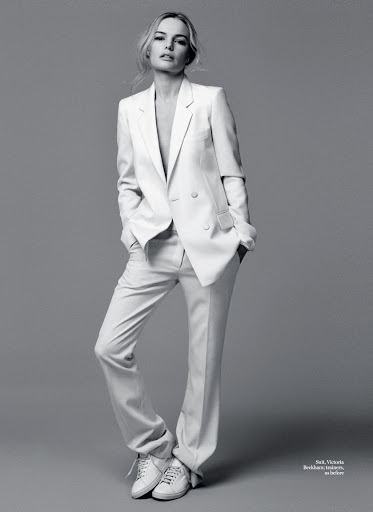 Monochrome portrait of a woman wearing a white suit jacket, matching trousers, and white trainers against a grey background.